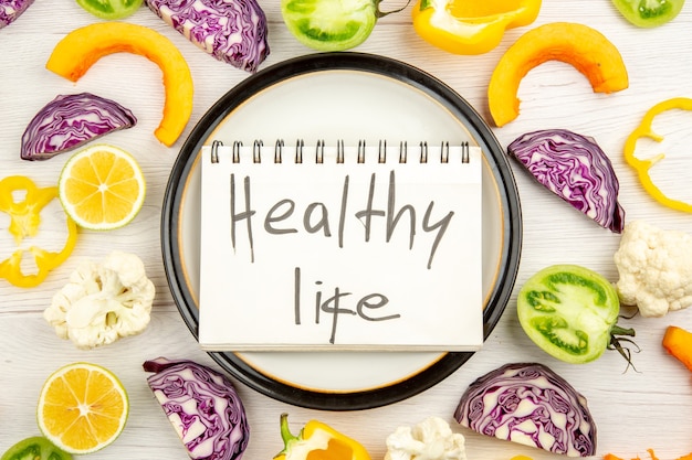 Top view healthy life written on notepad on round platter cut vegetables on white surface