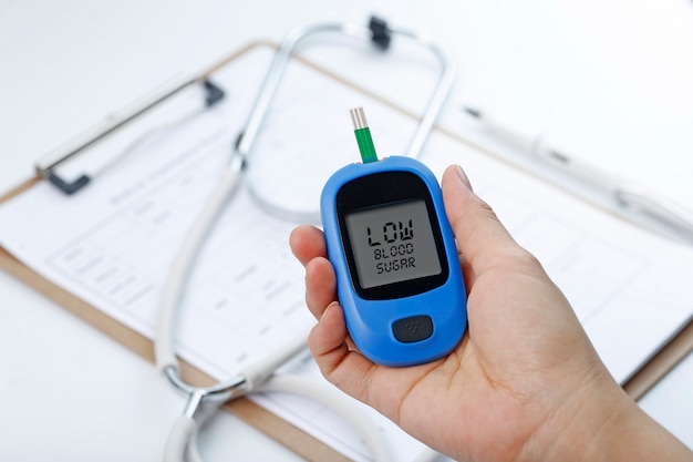 Hand holding a blood glucose meter measuring blood sugar, the background is a stethoscope and chart file